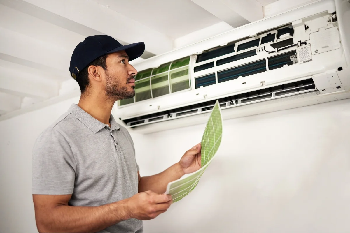 A man in a gray shirt and cap inspects and replaces the filters of a wall-mounted air conditioner, showcasing his expertise in HVAC emergency services.