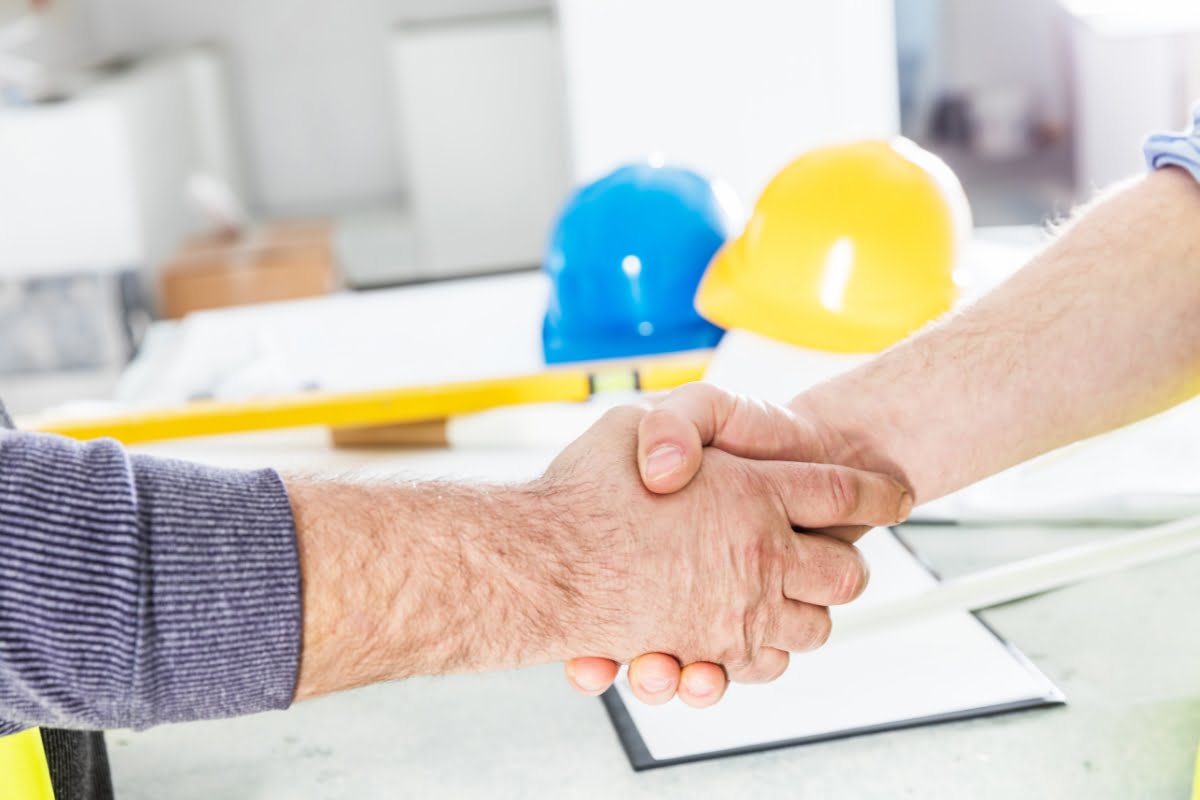 Two people shaking hands in an office setting, with construction helmets and blueprints visible in the background, suggesting a discussion involving hvac emergency services.