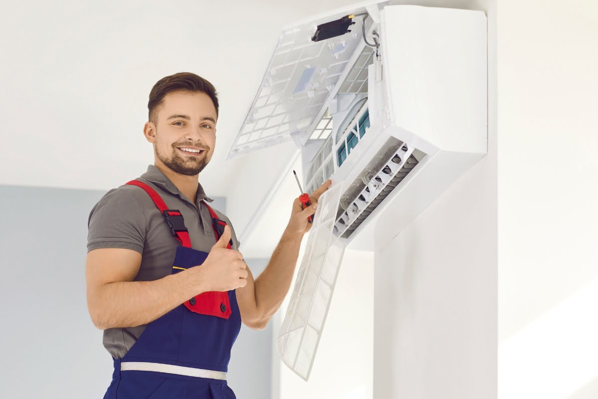 A technician in overalls holds a screwdriver and gives a thumbs-up while providing hvac emergency services on a wall-mounted air conditioning unit.