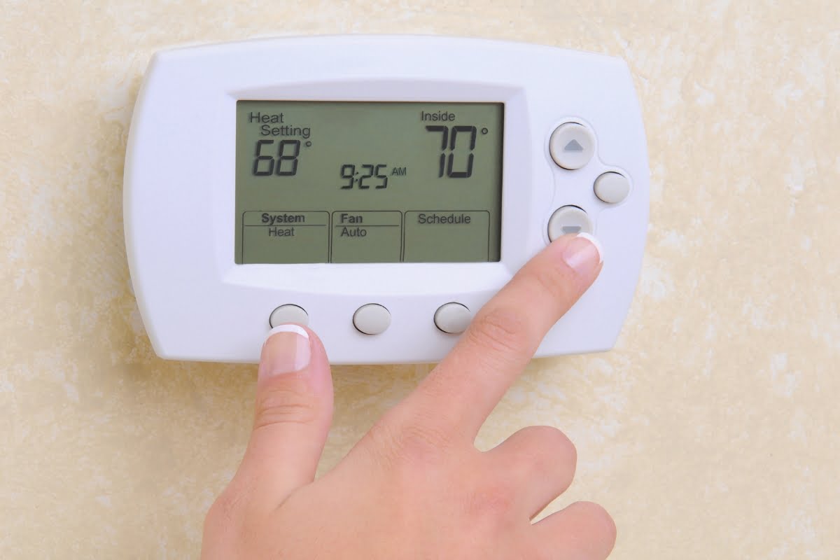 A hand adjusts a wall-mounted digital thermostat, part of advanced HVAC energy solutions. The display shows a heat setting of 68°F, an inside temperature of 70°F, and a time of 9:25 AM. Four buttons and a directional pad are visible.