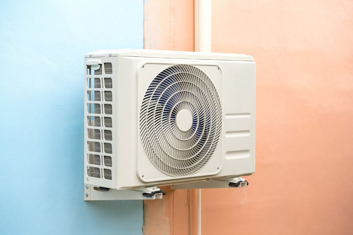 A white outdoor air conditioning unit, reflecting advanced HVAC energy solutions, is mounted on a wall painted half blue and half peach.