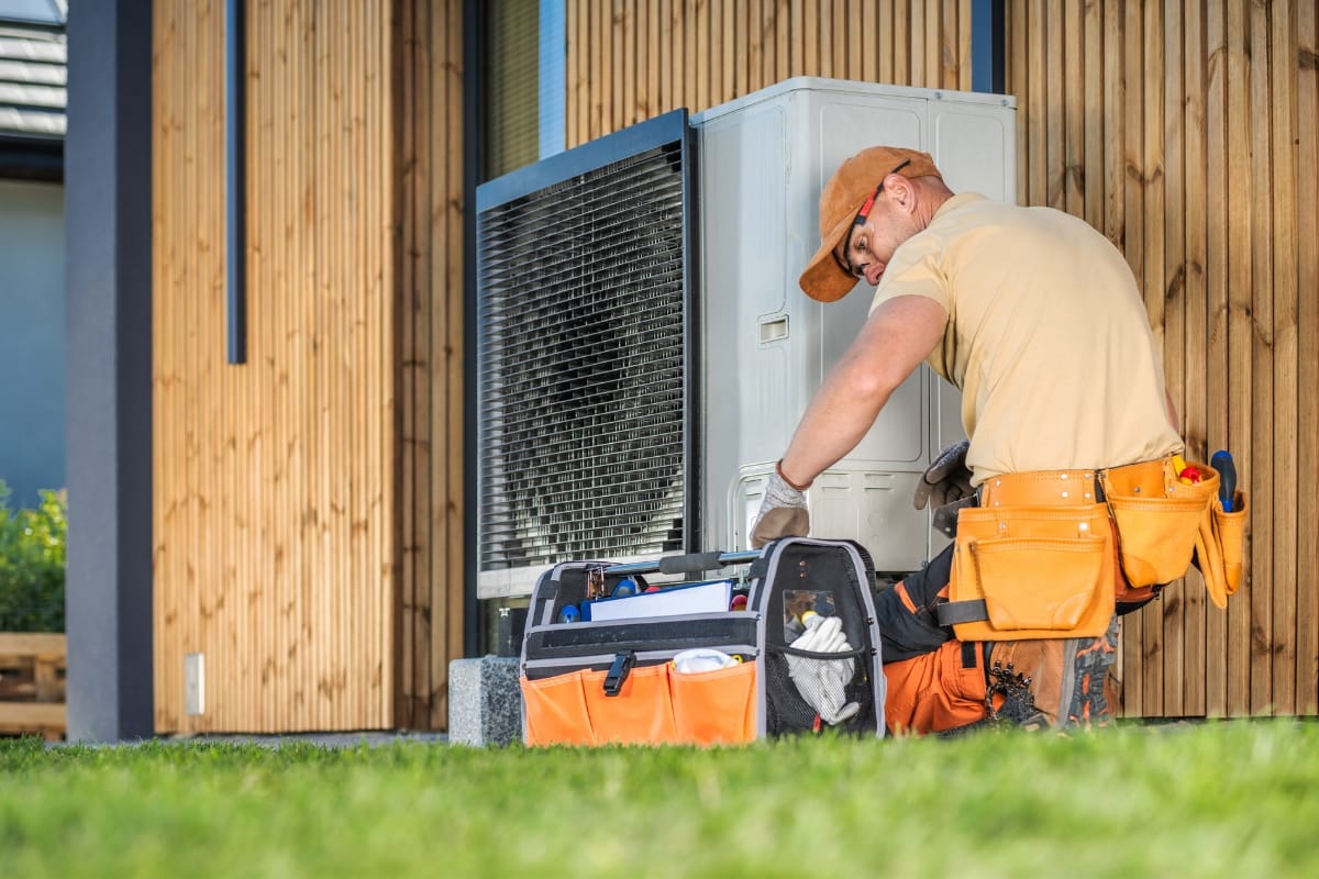 A technician in an orange cap and tool belt is performing an HVAC inspection on an outdoor air conditioning unit next to a wooden wall. A tool bag is open on the grass beside him.