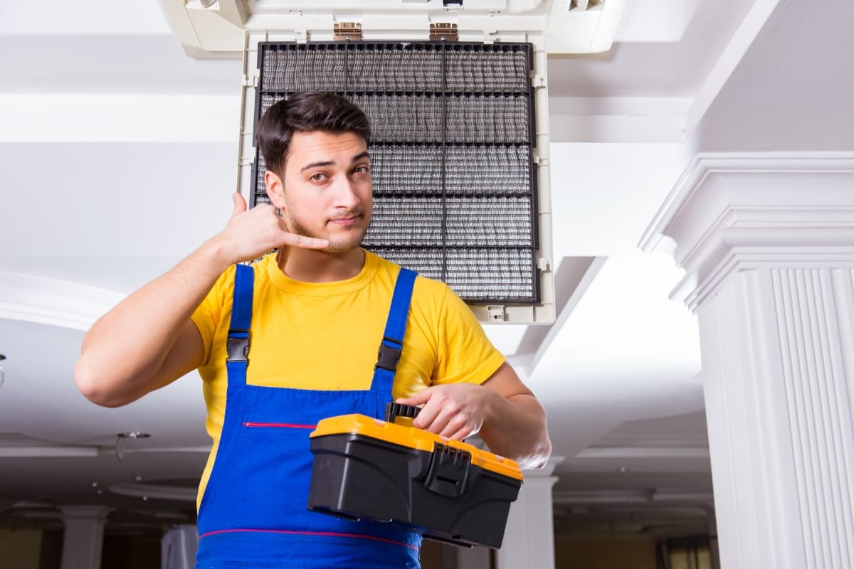 A man in a blue overall and yellow shirt stands under an open ceiling vent, holding a toolbox. He gestures with his hand near his ear, clearly well-versed in different types of HVAC systems.