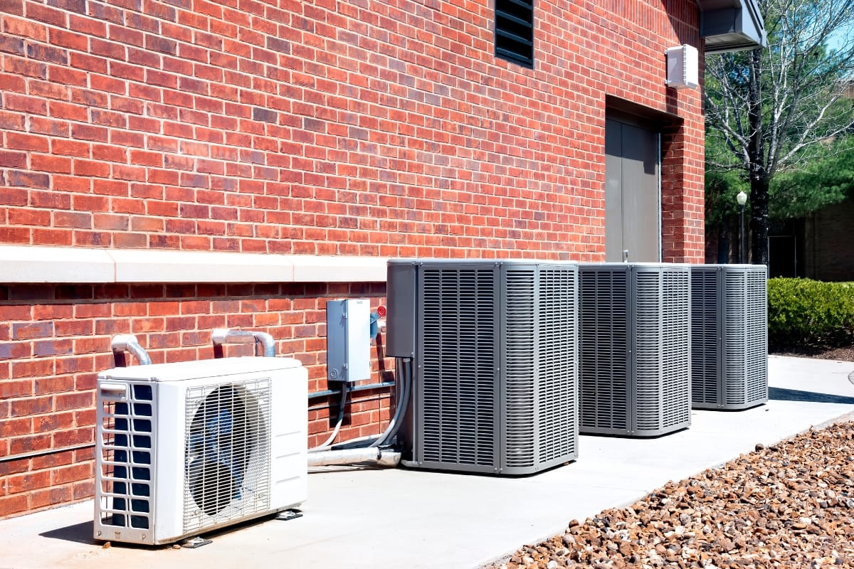 Four outdoor air conditioning units, representing different types of HVAC systems, are placed on a concrete slab next to a brick building. Nearby, gravel and a tree add to the serene setting.