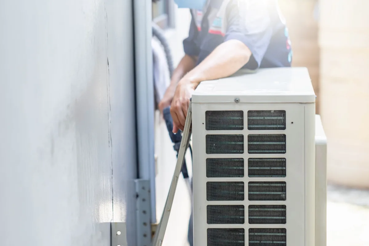 A technician installs or services an outdoor air conditioning unit mounted on a wall, ensuring it's compatible with various types of HVAC systems.