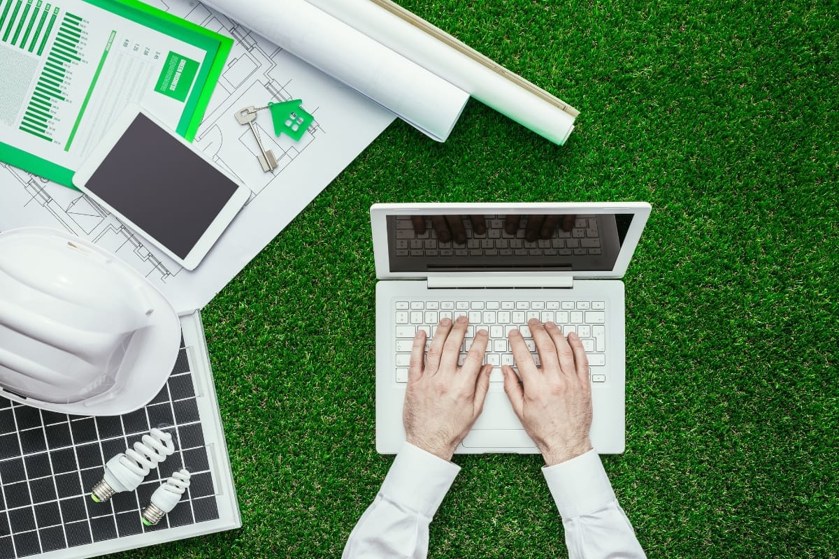 A person types on a laptop placed on grass, surrounded by blueprints, a tablet, a hard hat, CFL bulbs, and sustainability charts—an emblem of environmental conservation in action.