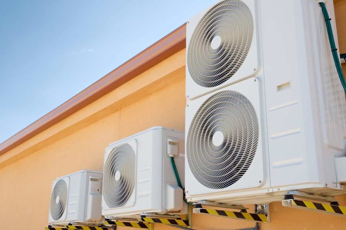 Three outdoor air conditioning units are mounted on a beige wall, under a clear blue sky—a stark reminder of the need for environmental conservation.