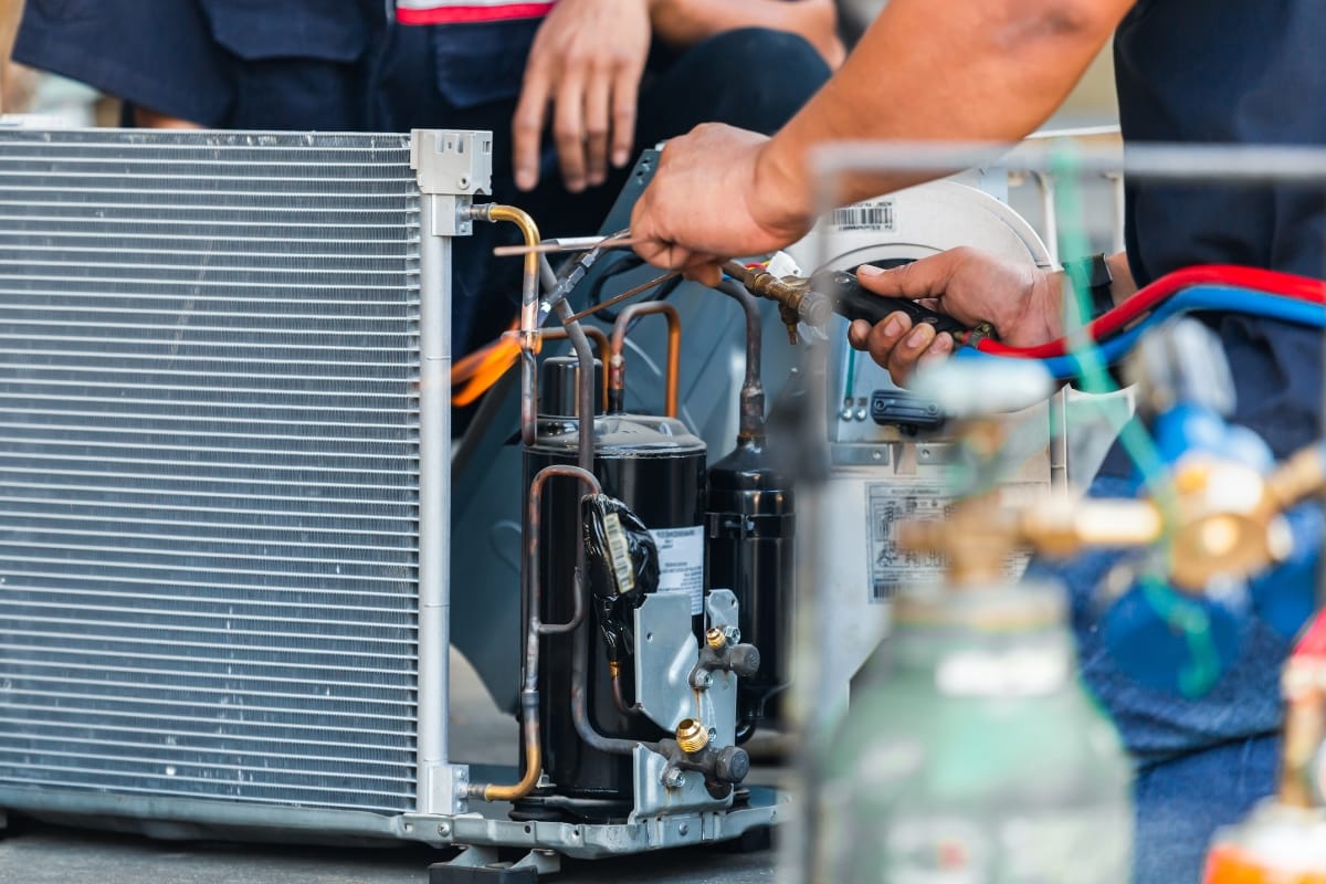 Technicians meticulously repairing an air conditioning unit, employing tools and air pressure hoses, as visible components like copper pipes and cooling fins are examined—highlighting their expertise comparable to addressing the intricate cost of furnace repairs.