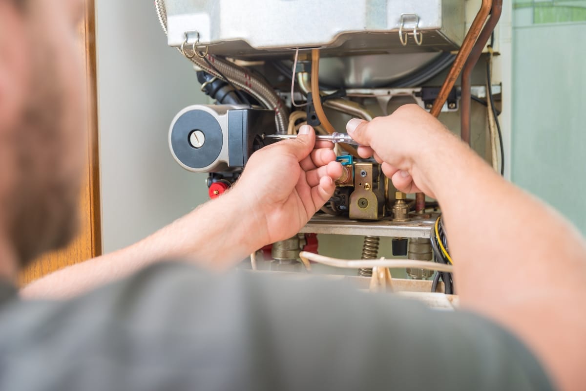A person uses a wrench to repair a gas boiler, tackling what appears to be an emergency furnace repair amidst various wires and components.