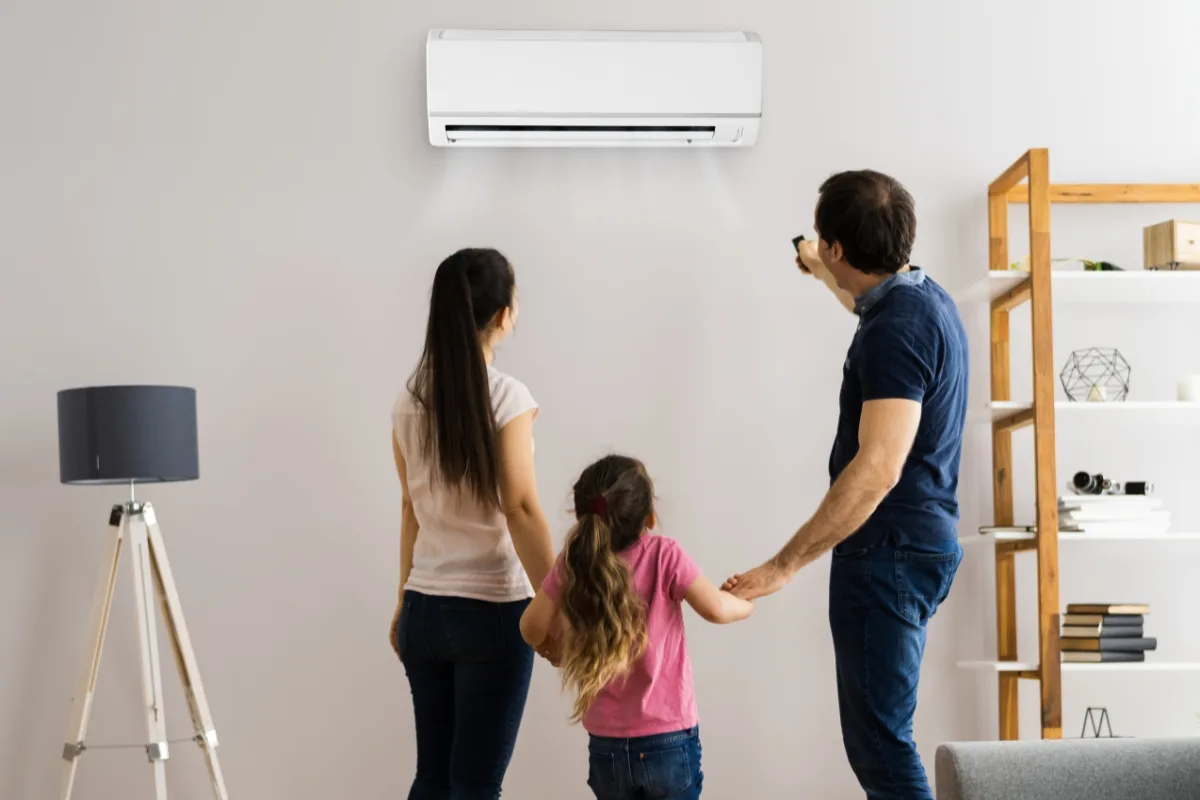 In their cozy Holly Springs living room, a family of three stands before an air conditioner. The man aims the remote, determined to fix any AC problems that arise. Shelving and a lamp enhance the charming backdrop.