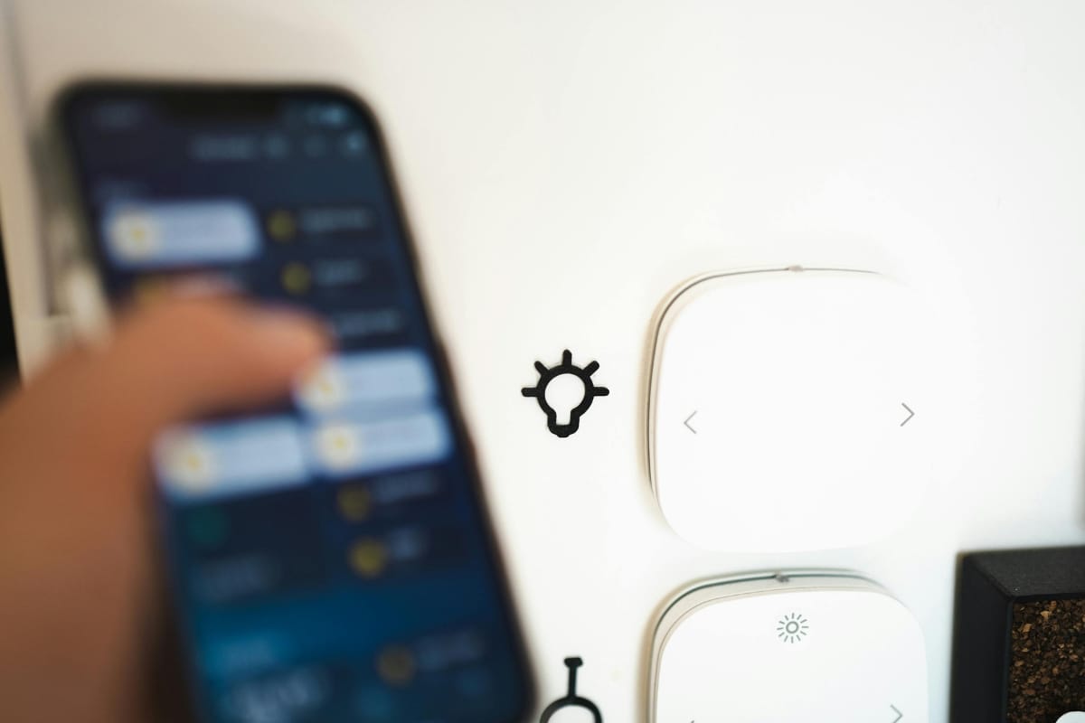 A person holds a smartphone displaying a blurred app screen near a wall with two white smart home devices, showcasing smart thermostat benefits, and a black gear-shaped wall hook.