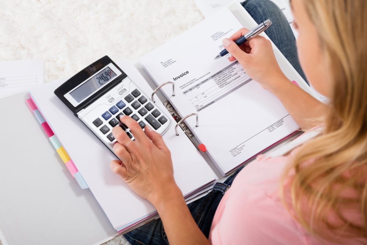 Person using a calculator and reviewing an invoice in a binder with colored tabs, possibly noting smart thermostat benefits for energy savings.