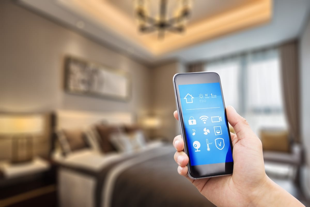 A hand holds a smartphone displaying a smart home control app in a modern, well-lit bedroom with a large bed and soft lighting, highlighting smart thermostat benefits for personalized comfort and energy efficiency.