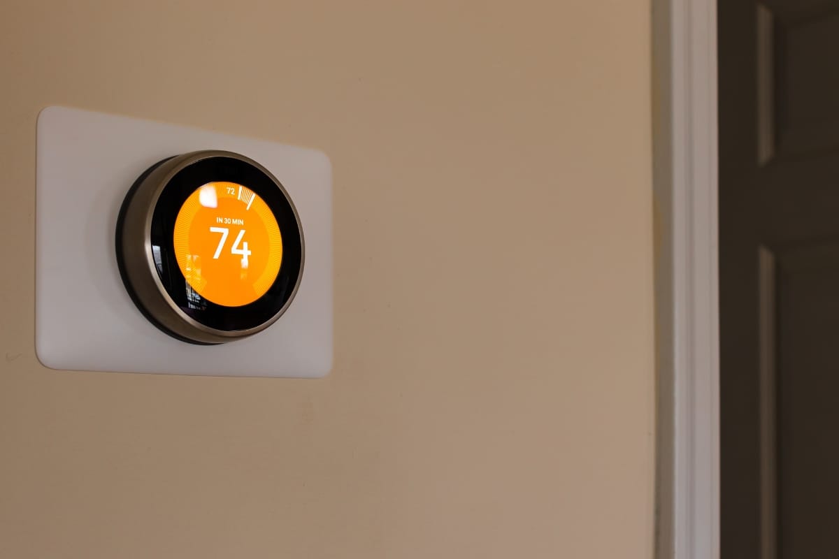 A smart thermostat displays a temperature of 74 degrees on a beige wall next to a white door frame, showcasing smart thermostat benefits like easy monitoring and energy efficiency.
