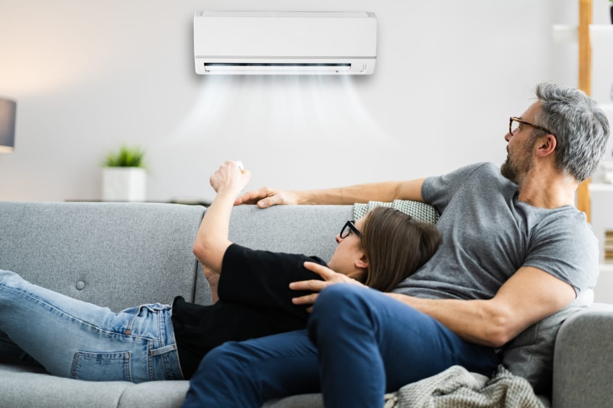 Two people relax on a couch, one using a remote to control the best air conditioner mounted on the wall, enjoying the cool air flowing into the room.