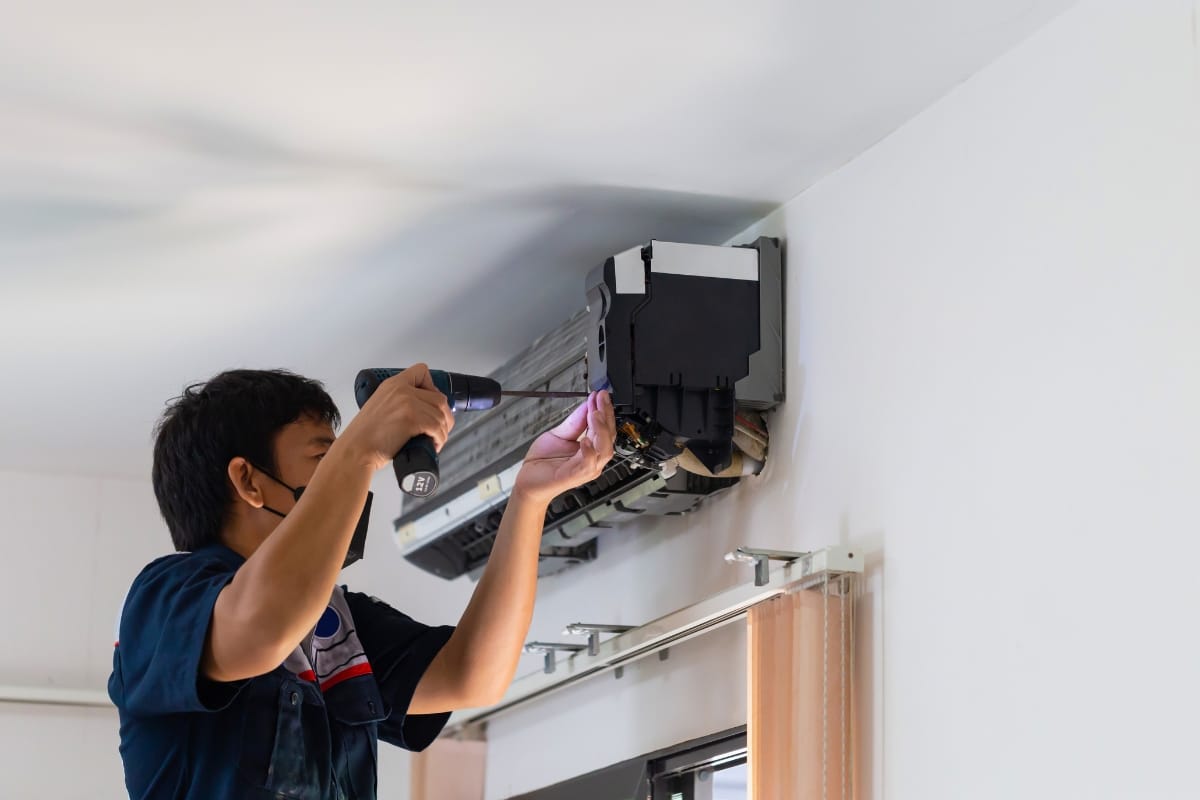 A technician wearing a mask uses a power drill to repair one of the best air conditioners mounted on a white wall.