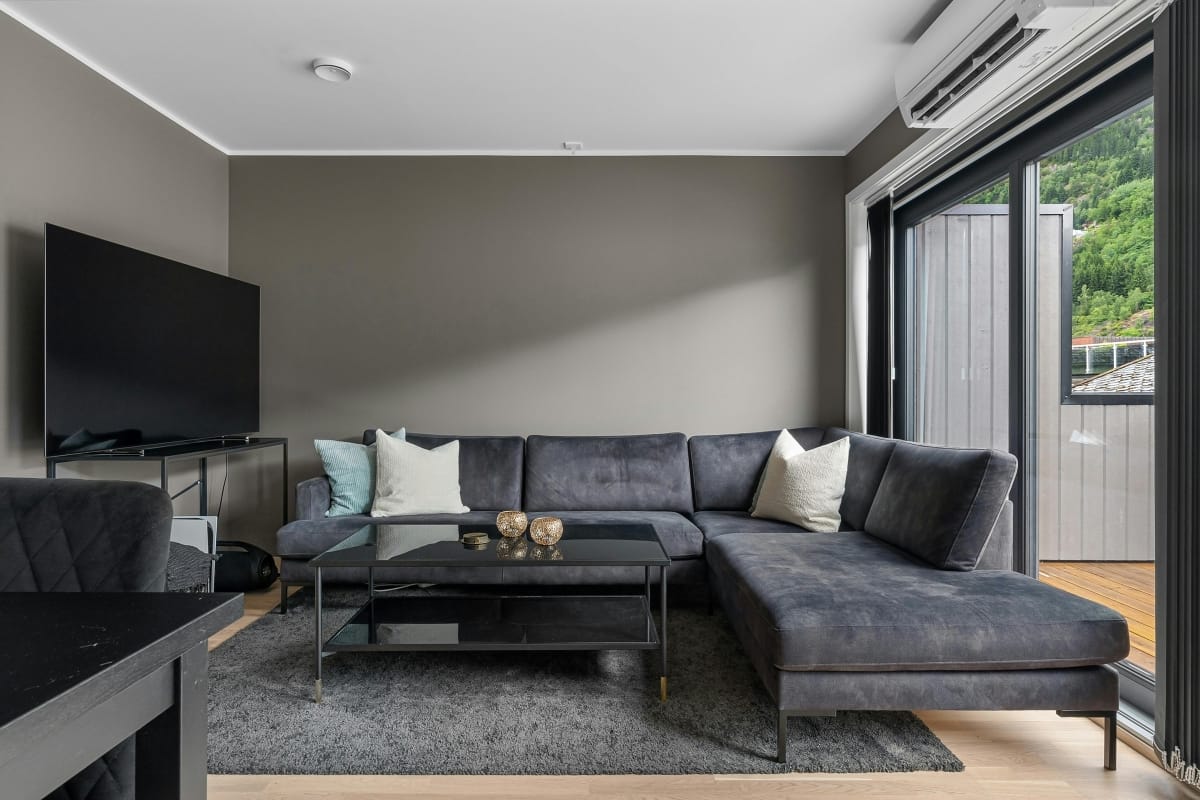 Modern living room with a dark gray sectional sofa, glass coffee table, large TV, and sliding glass door opening to a balcony with a view of greenery—kept cool year-round with one of the best air conditioners.
