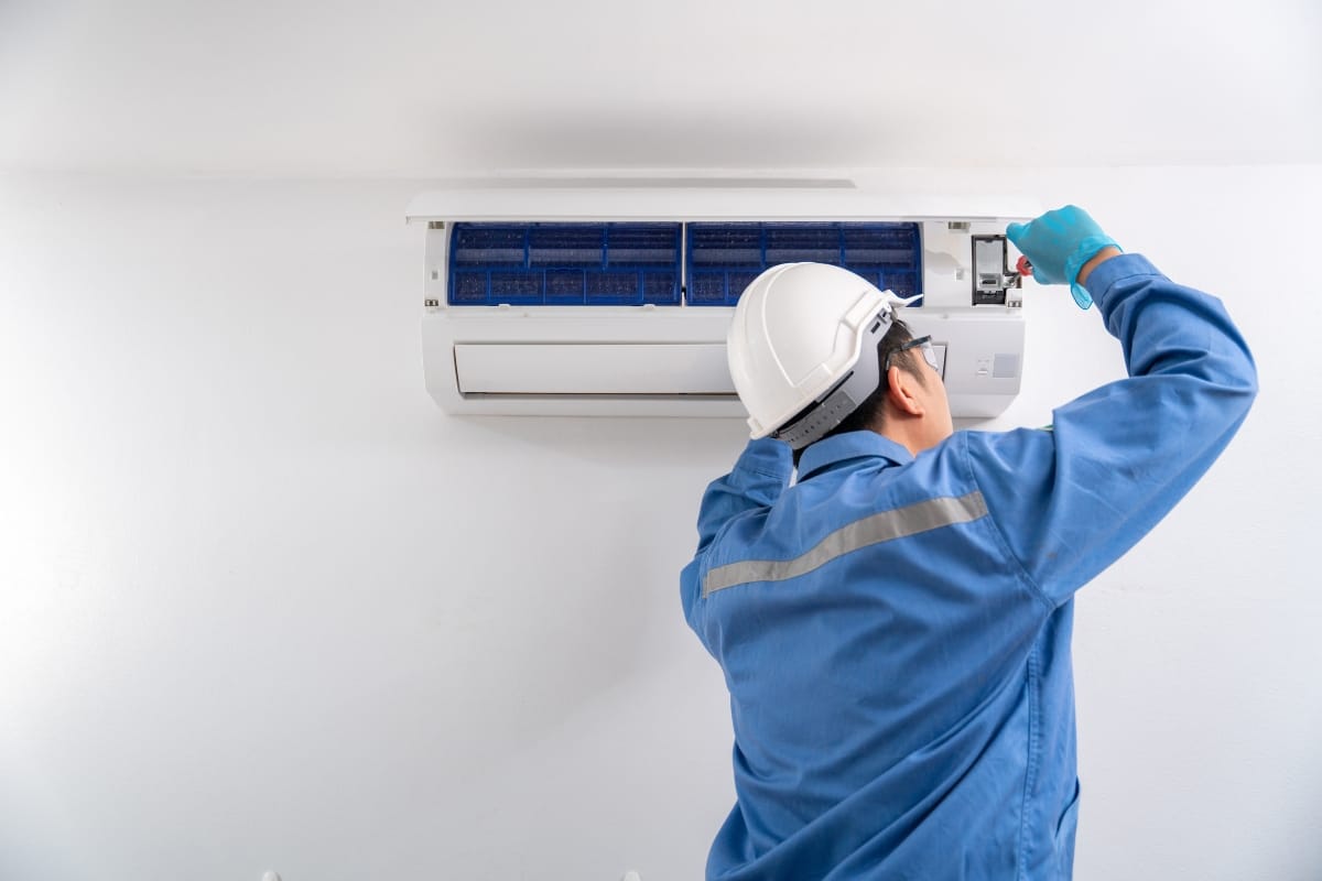 A technician in a blue uniform and hard hat performs an emergency AC repair as he inspects or repairs a wall-mounted air conditioning unit.