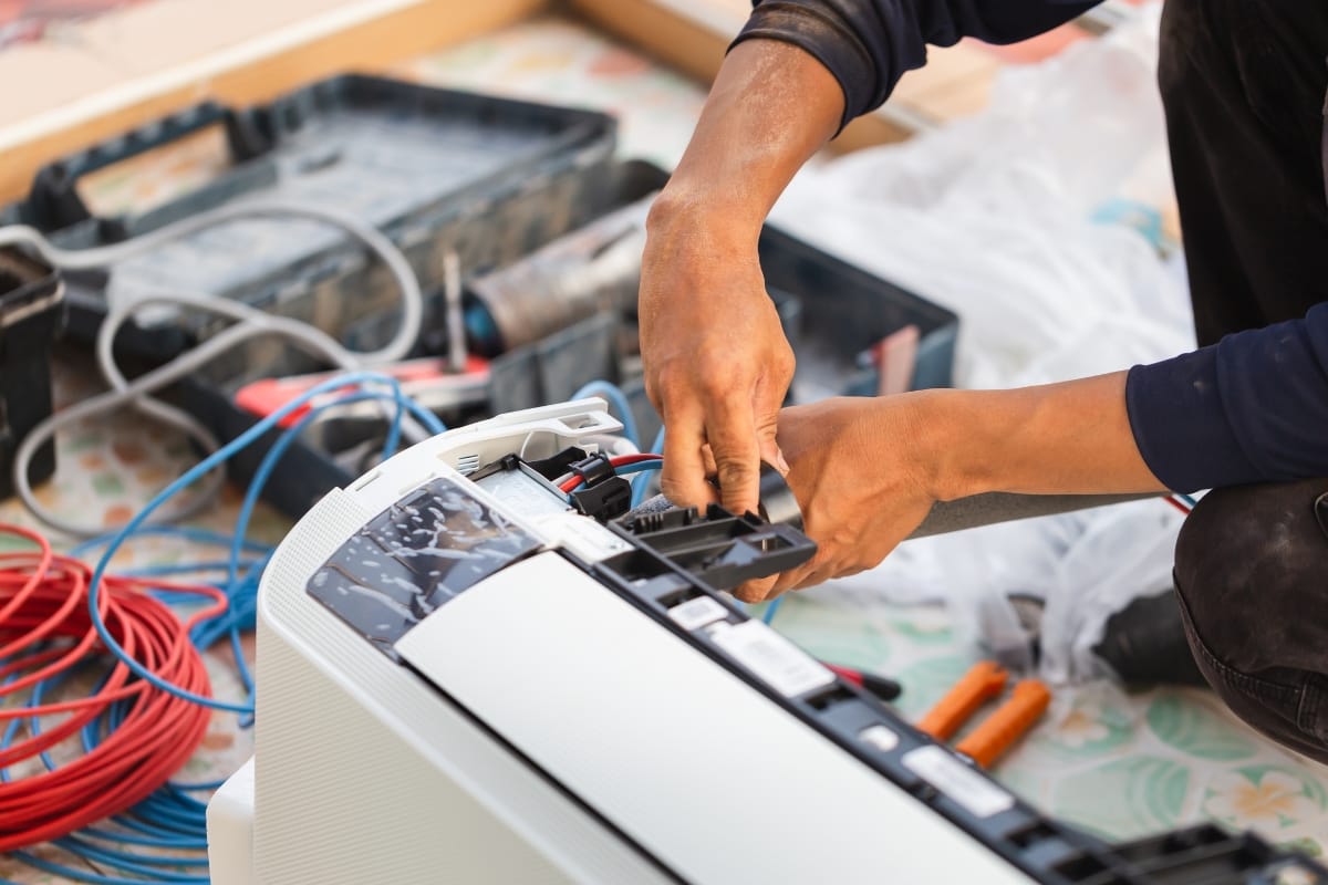A person performs emergency AC repair, installing or fixing an air conditioning unit with tools and wires, while a toolbox and equipment are visible in the background.