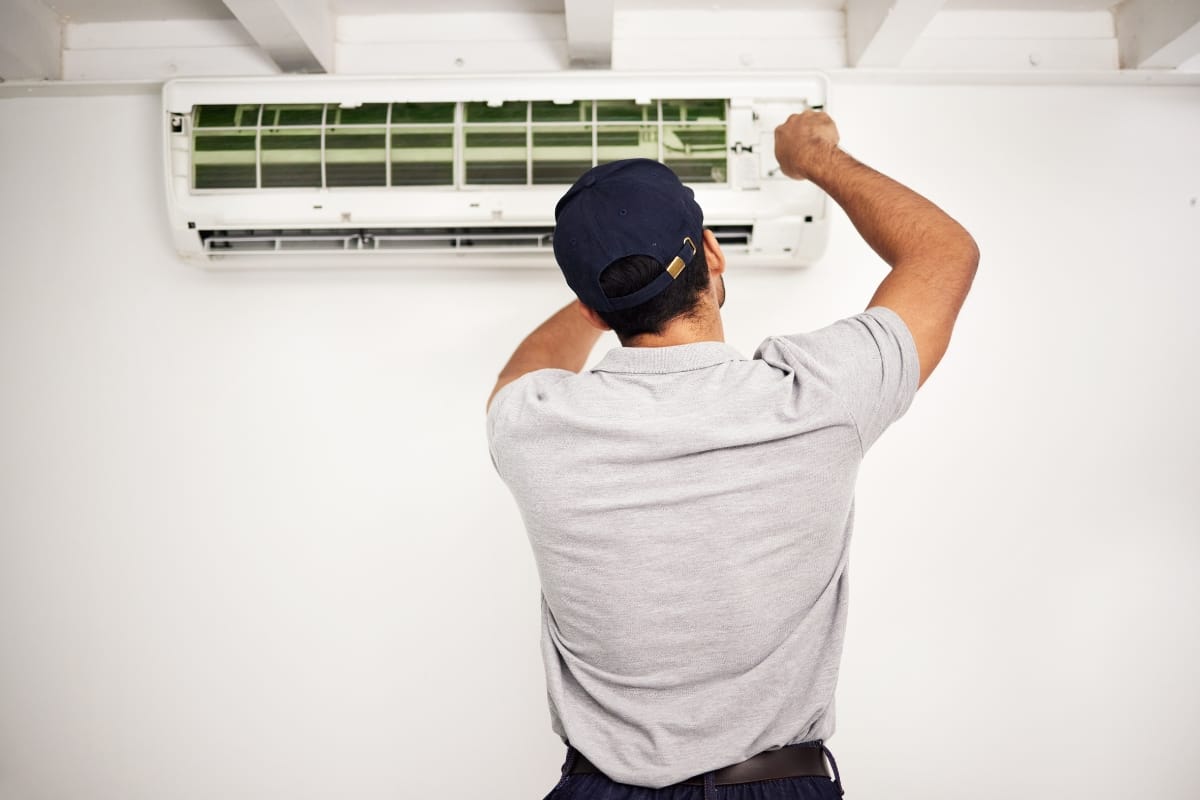 A technician in a gray shirt and navy cap is performing an Emergency AC Repair, inspecting or fixing a wall-mounted air conditioning unit.