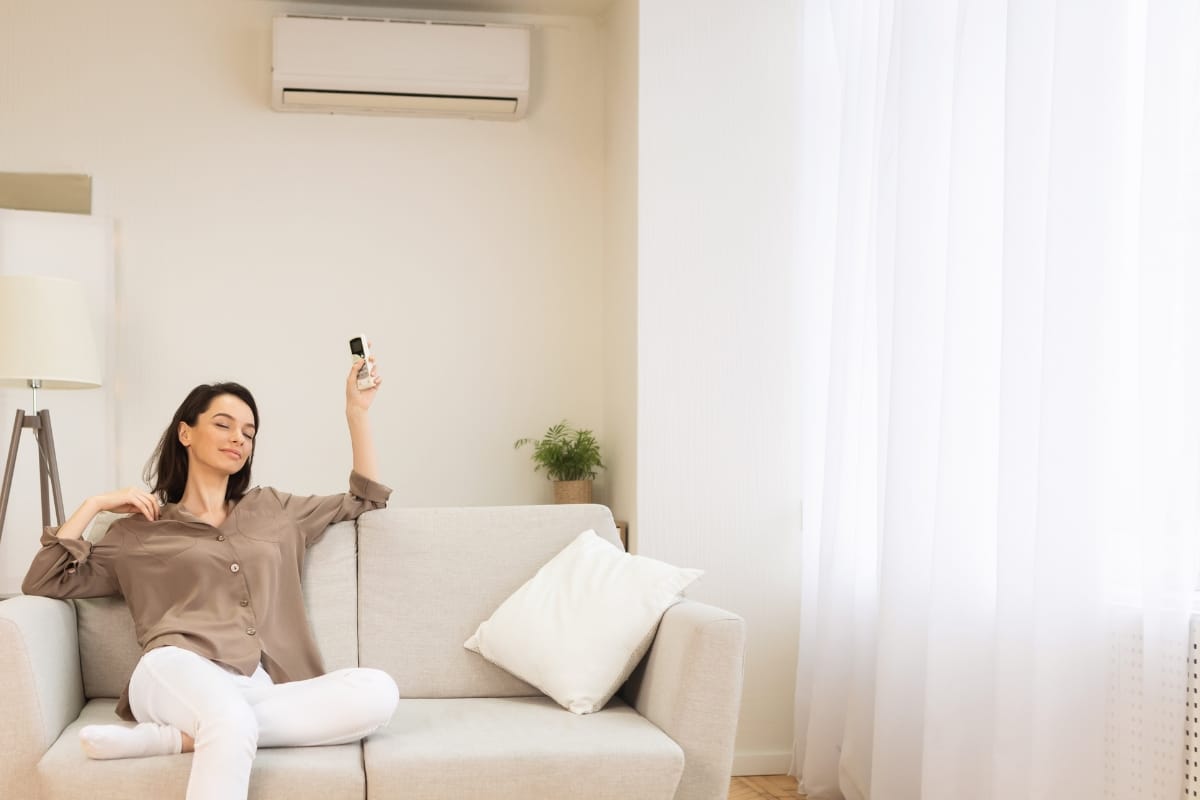 Woman sitting on a sofa using a remote control to operate an air conditioner in a bright living room with white curtains, feeling confident knowing Emergency AC Repair is just a call away if needed.