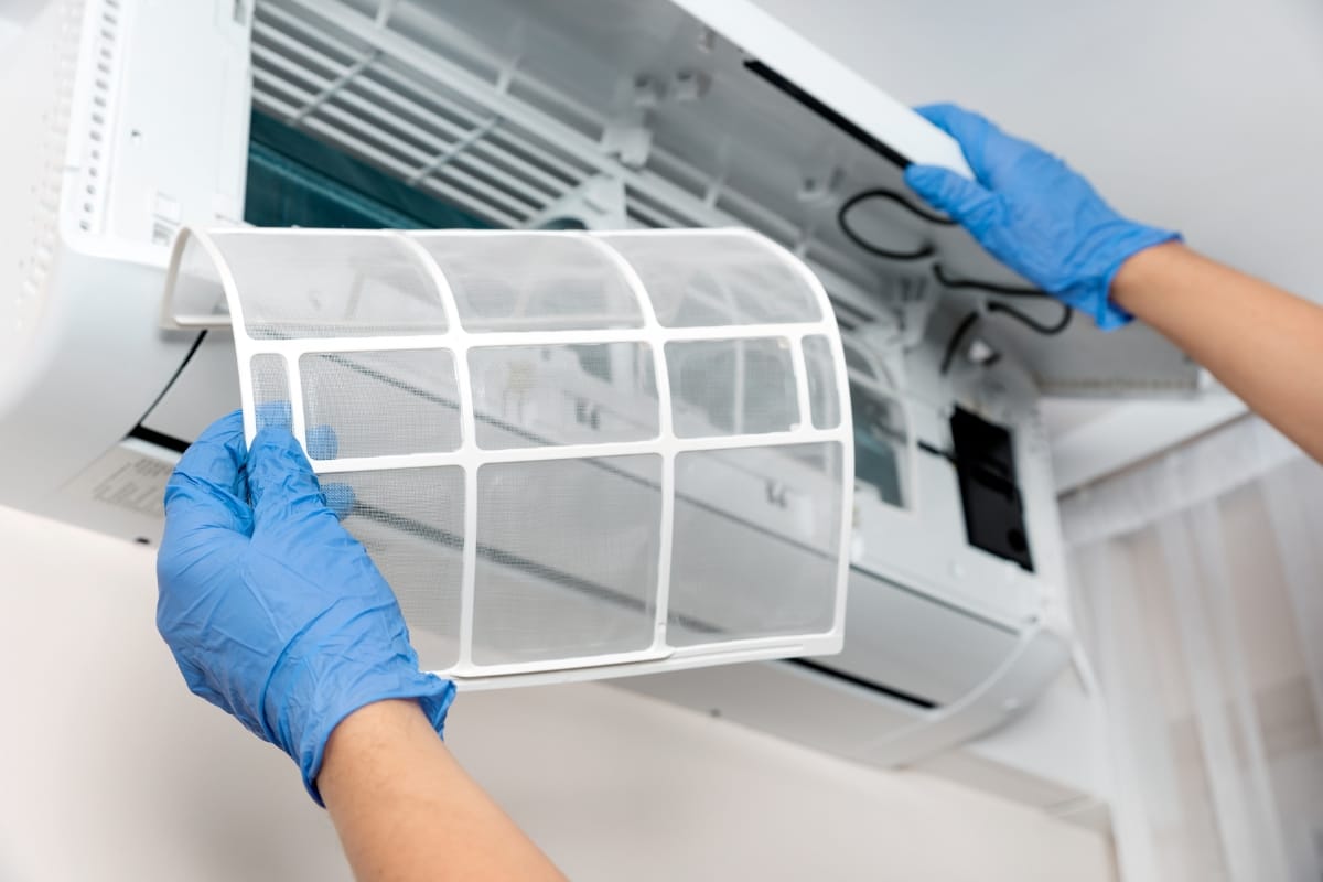 A person wearing blue gloves removes a mesh filter from a wall-mounted air conditioner for cleaning or maintenance, an important step in preventing the need for emergency AC repair.