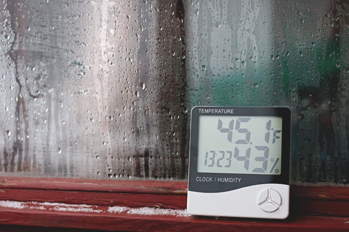 A digital thermometer and hygrometer displays 45.1°F and 34% humidity, placed on a red windowsill beside a condensation-covered window—ideal for monitoring effective humidity control indoors.