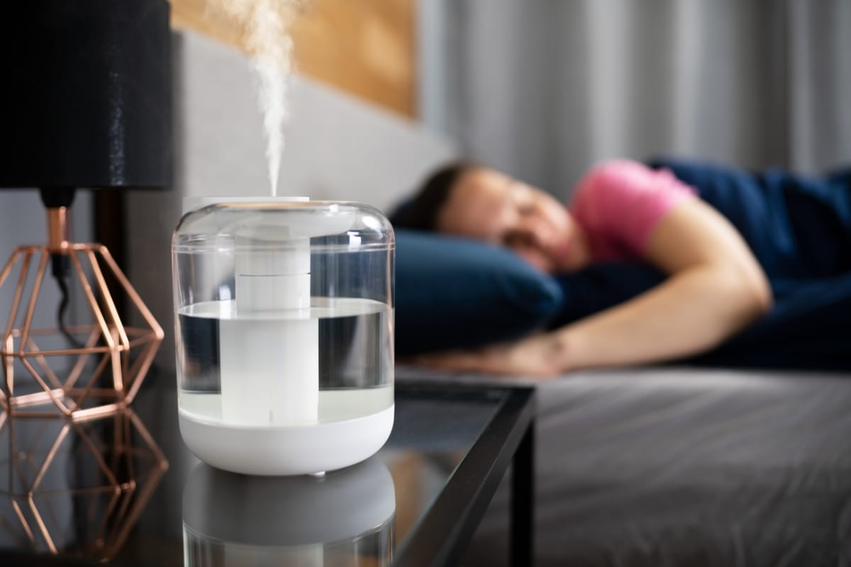 A clear humidifier with advanced humidity control emits mist on a bedside table, while a person sleeps peacefully in the background.