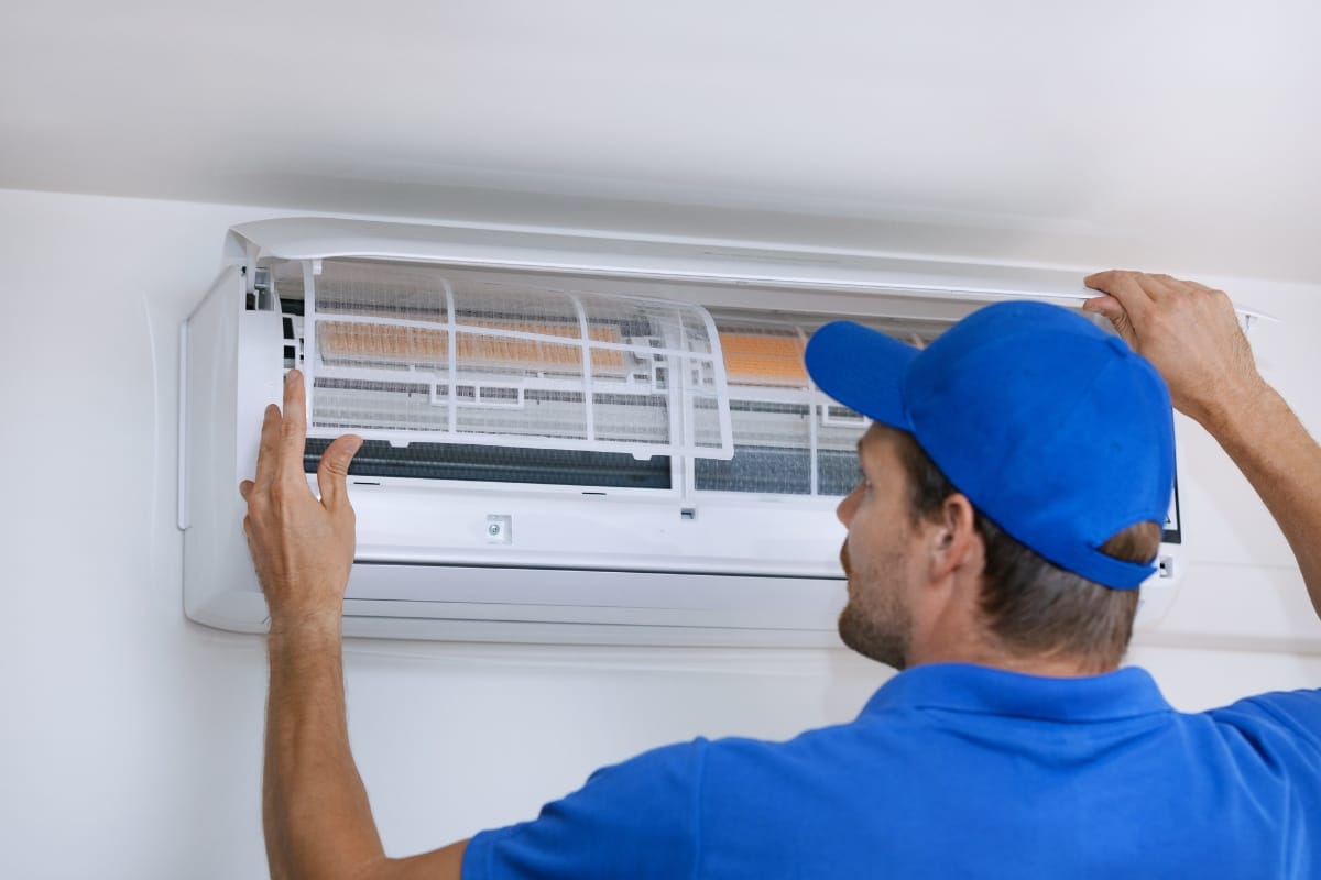 A technician in a blue uniform from All-Season Heating & Cooling inspects or repairs a wall-mounted air conditioning unit indoors.