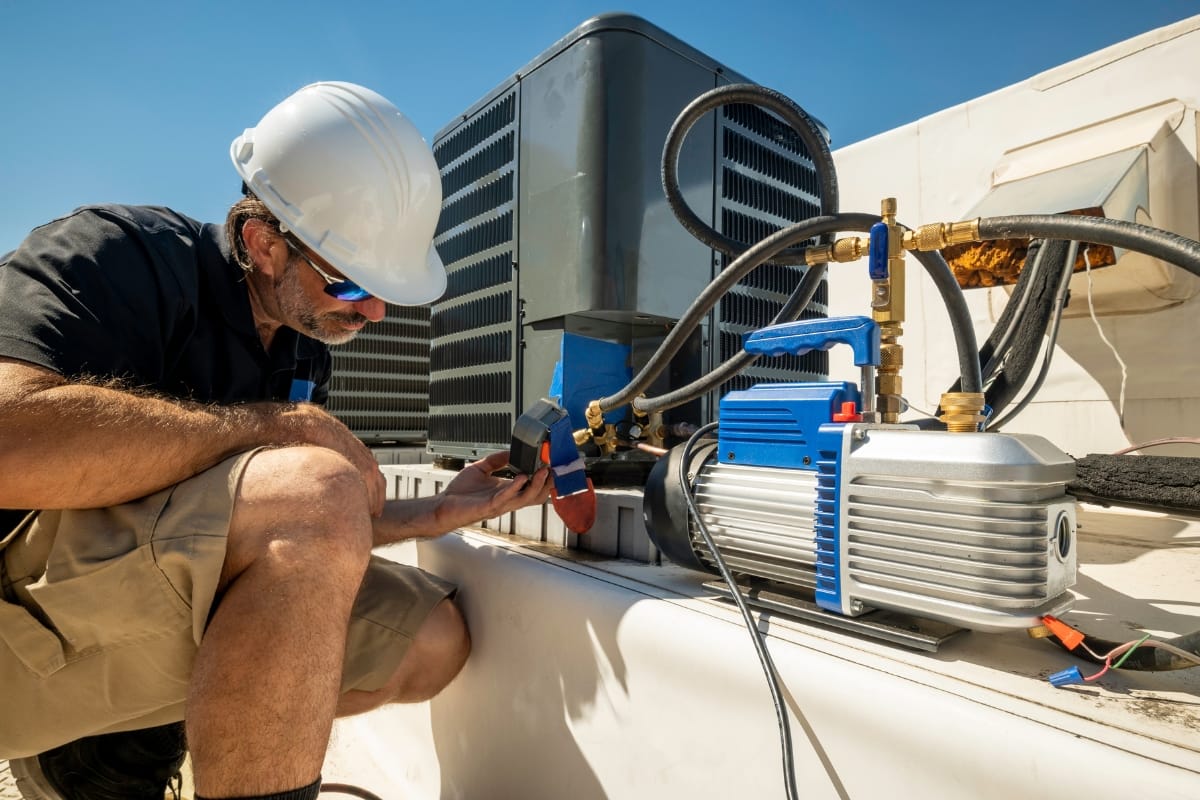 A technician from All-Season Heating & Cooling, wearing a hard hat, kneels beside an outdoor HVAC unit, using tools and gauges to perform maintenance or repairs.