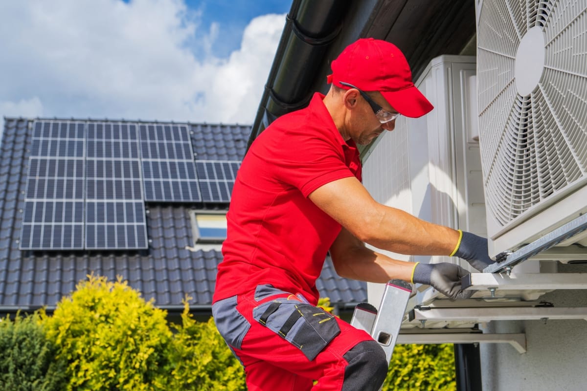 A technician in red uniform installs or services an outdoor air conditioning unit on a house with solar panels, representing All-Season Heating & Cooling’s commitment to efficient, sustainable comfort year-round.