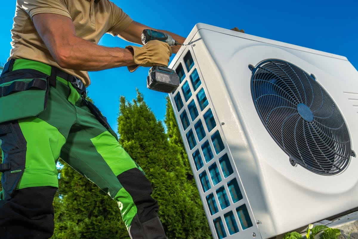 A technician in green work pants uses a power drill to install or repair an outdoor All-Season Heating & Cooling unit beside green trees under a clear blue sky.