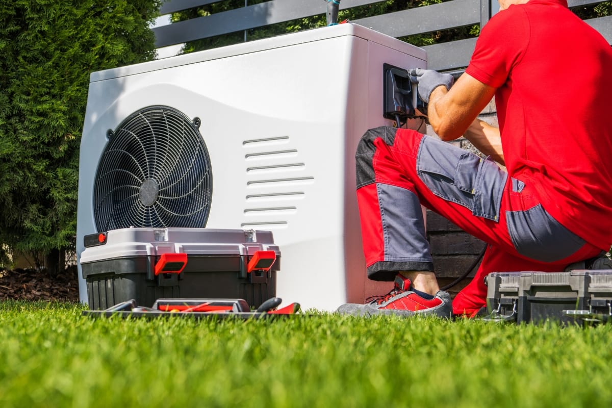A technician in red attire from All-Season Heating & Cooling services an outdoor heat pump unit, with tools and toolboxes scattered on the grass nearby.