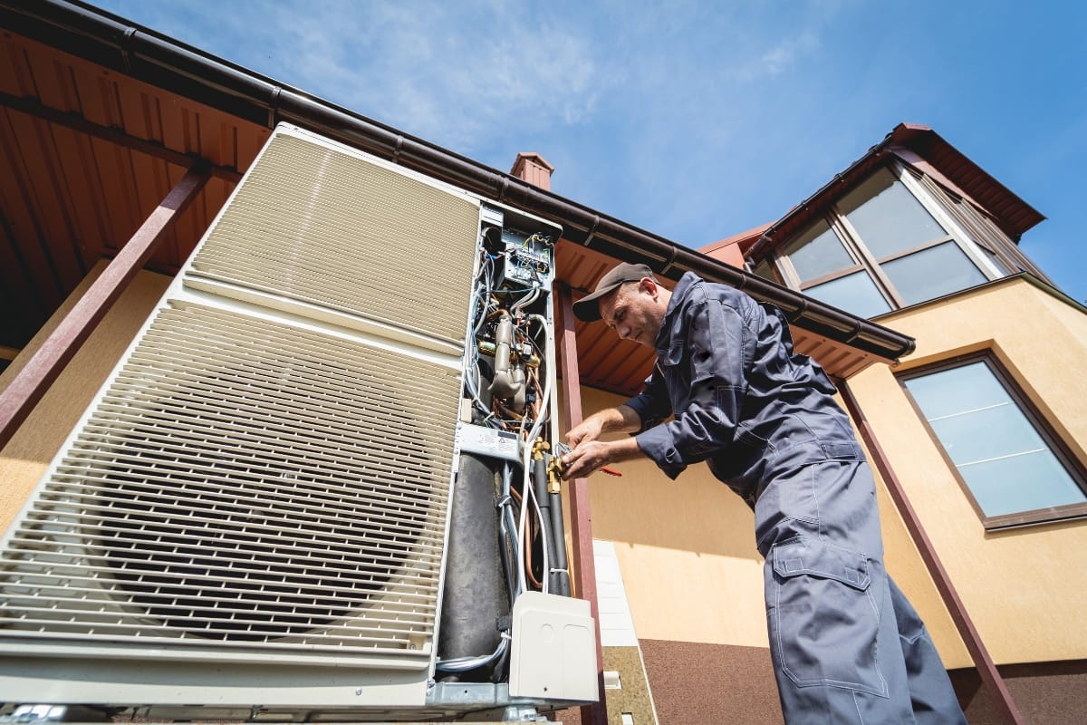 A technician in uniform repairs an outdoor air conditioning unit next to a house under a clear blue sky, ensuring the HVAC system is ready for the back-to-school season.
