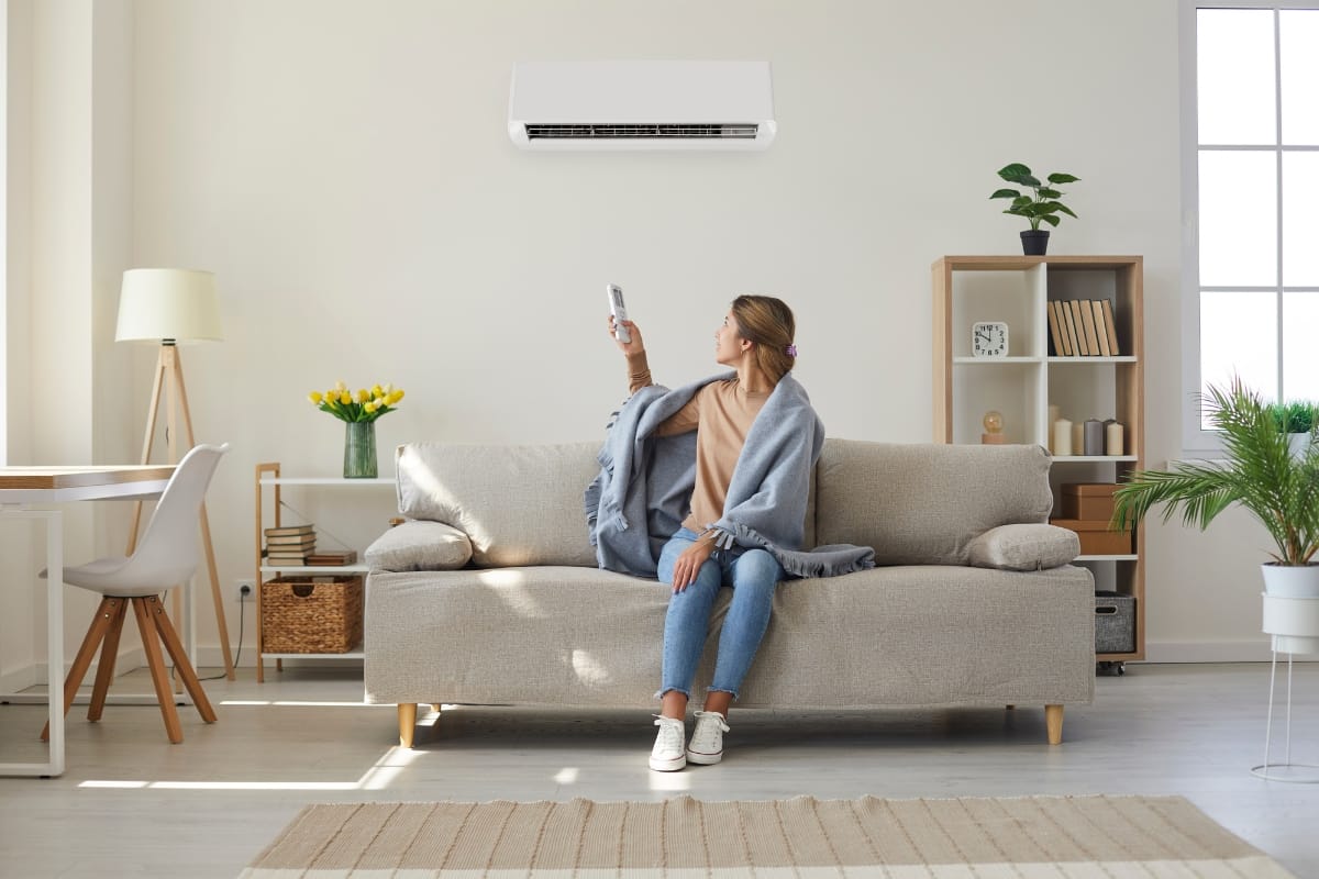 A woman sits on a sofa in a living room, covered with a blanket, using a remote control to operate the back-to-school HVAC system mounted on the wall above her.