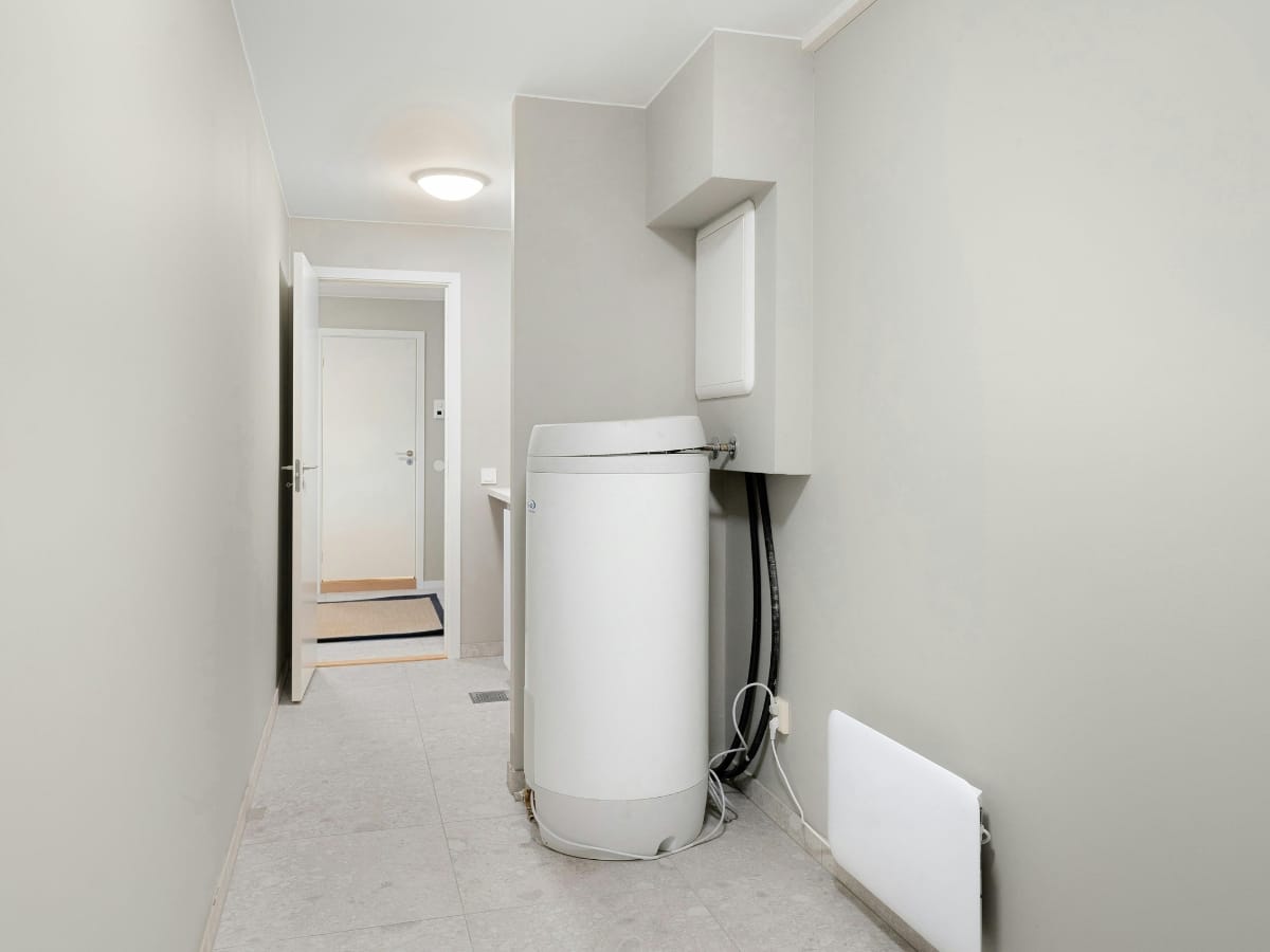 A laundry room with light gray walls, a white washing machine, wall-mounted utility boxes, tile flooring, and an open door leading to a hallway—ideal for homes using the best heating systems for year-round comfort.