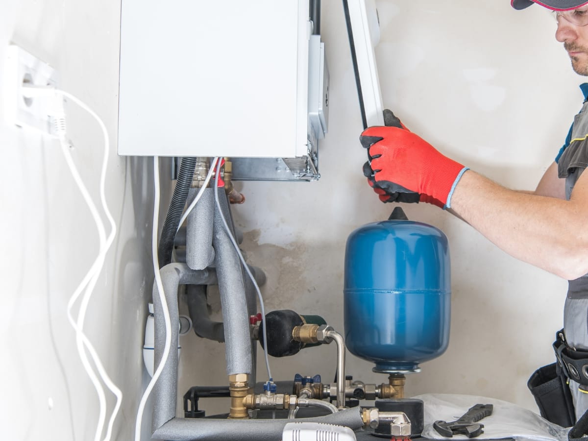 A worker in red gloves is adjusting a blue expansion tank connected to a wall-mounted boiler system, showcasing features of the Best Heating Systems for efficient home comfort.