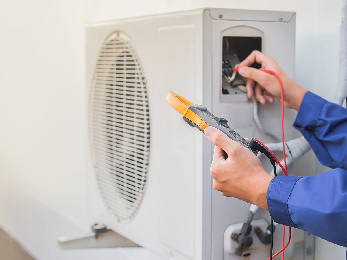A person in a blue uniform uses a digital multimeter to check the electrical connections of an outdoor air conditioning unit, ensuring optimal performance alongside the best heating systems.
