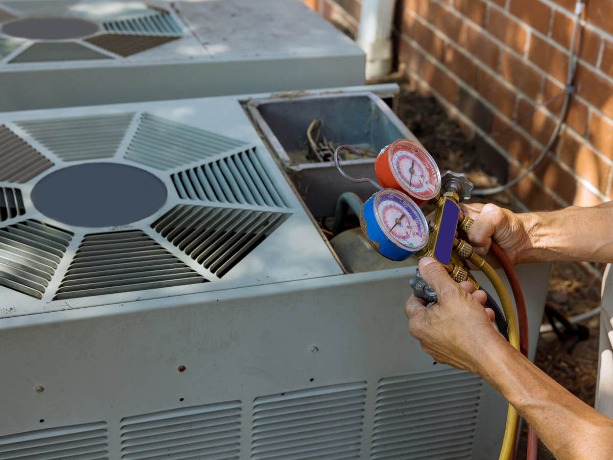 A person uses a manifold gauge set to check the refrigerant levels on an outdoor air conditioning unit near a brick wall as part of their Fall HVAC Maintenance routine.