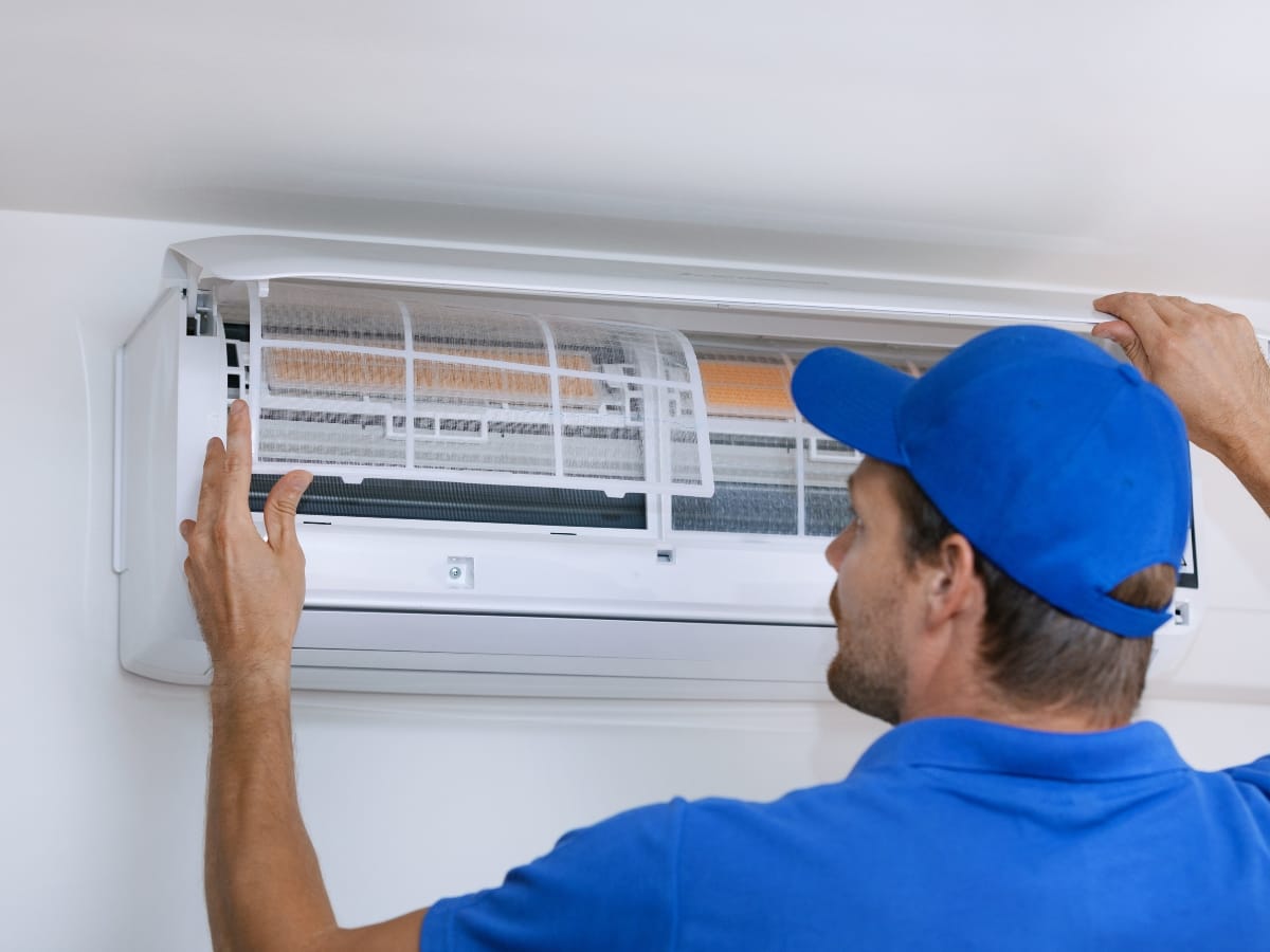 A technician wearing a blue cap inspects or repairs an open wall-mounted air conditioning unit indoors as part of essential Fall HVAC Maintenance.