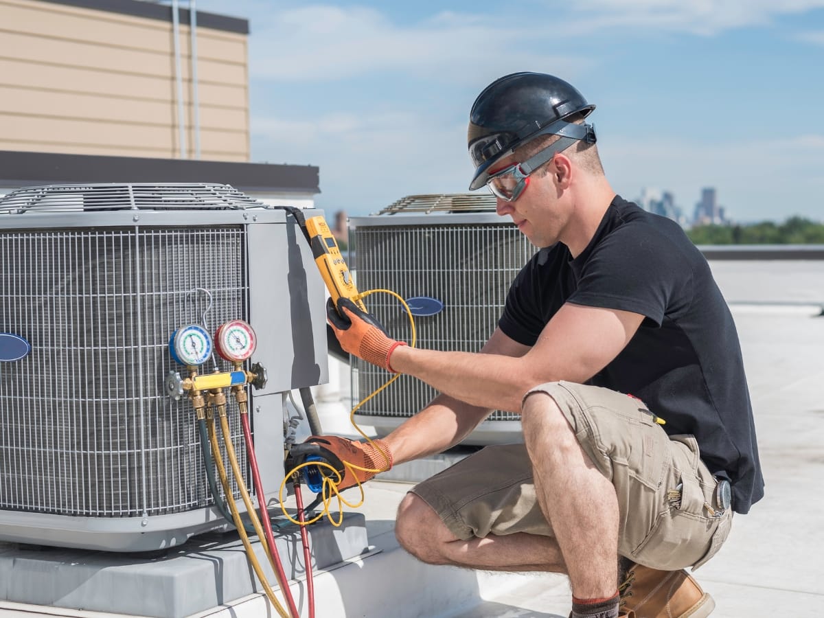 A technician wearing safety gear performs Fall HVAC Maintenance, using a multimeter to check an air conditioning unit's electrical connections on a rooftop.