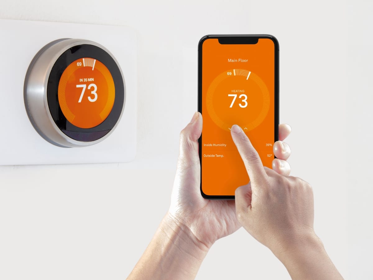 A person adjusts a smart thermostat with their smartphone, both displaying the temperature set to 73 degrees—perfect for ensuring comfort during Fall HVAC Maintenance.