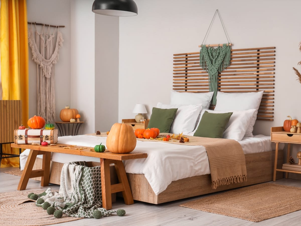 A modern bedroom decorated for autumn with pumpkins on the bed, bench, and nightstands, featuring green accents and cozy textiles—an inviting space perfect for relaxing after your seasonal fall HVAC maintenance.