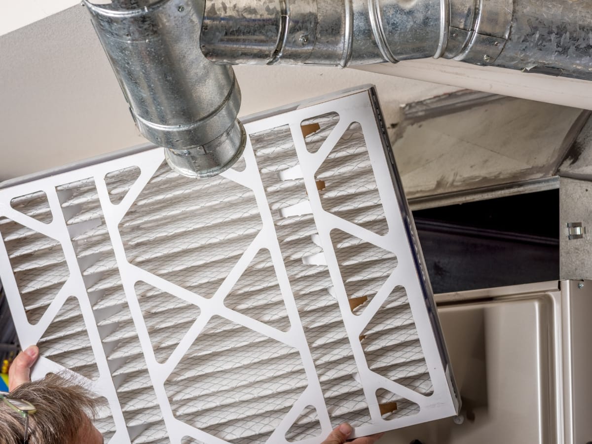 A person replaces a large HVAC air filter near an exposed duct and air vent in a ceiling—helping prevent one of the top heating problems caused by clogged or dirty filters.