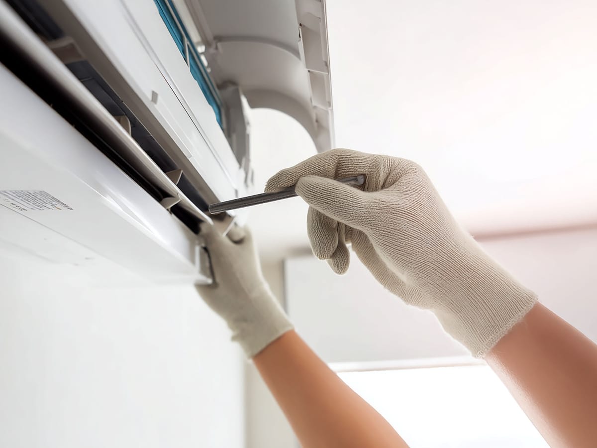 A person wearing gloves uses a screwdriver to service or repair an air conditioning unit mounted on a wall as part of essential Winter HVAC Maintenance.