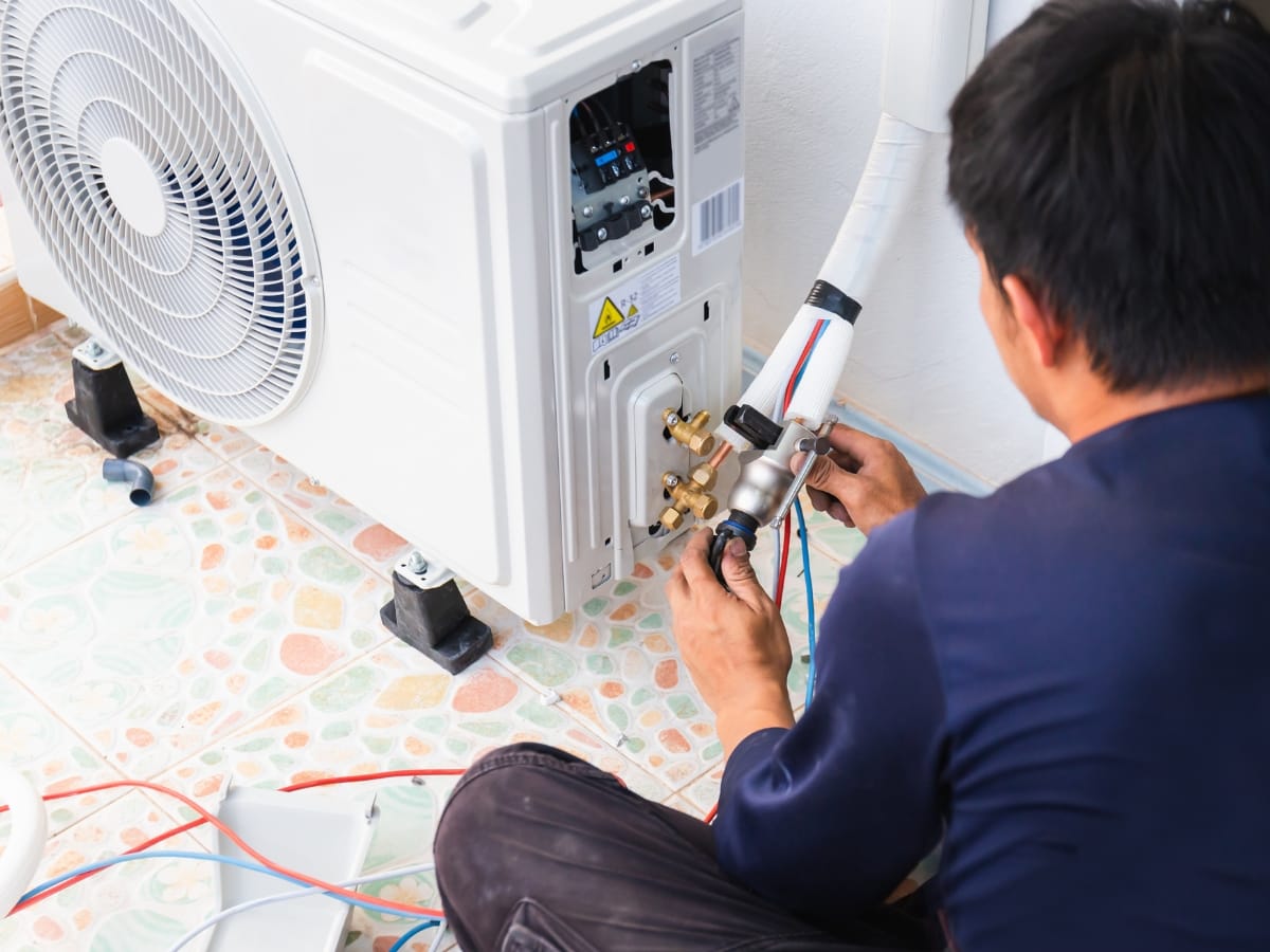 A technician performs Winter HVAC Maintenance, installing or repairing an outdoor air conditioning unit by connecting wires and pipes on a tiled floor.