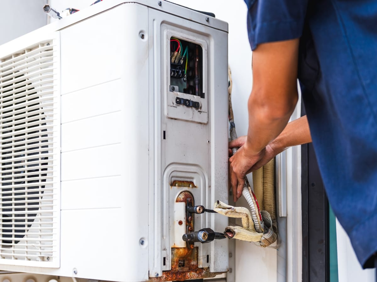 A person performs Winter HVAC Maintenance by repairing an outdoor air conditioning unit, with the cover open to expose wires and pipes.
