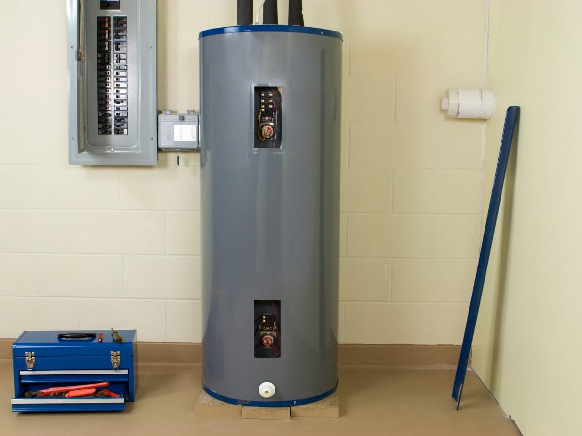 A tall, cylindrical water heater stands near an electrical panel and a blue toolbox in a utility room with cream walls and a beige floor, an ideal setting for scheduling your Annual Furnace Inspection.