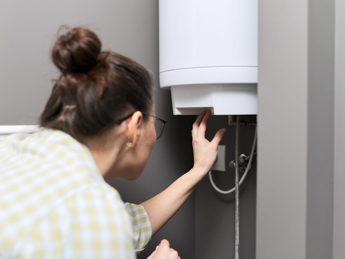A woman in glasses adjusts a control on a wall-mounted water heater with exposed pipes in a utility room, highlighting the importance of regular checks like an annual furnace inspection for home safety.