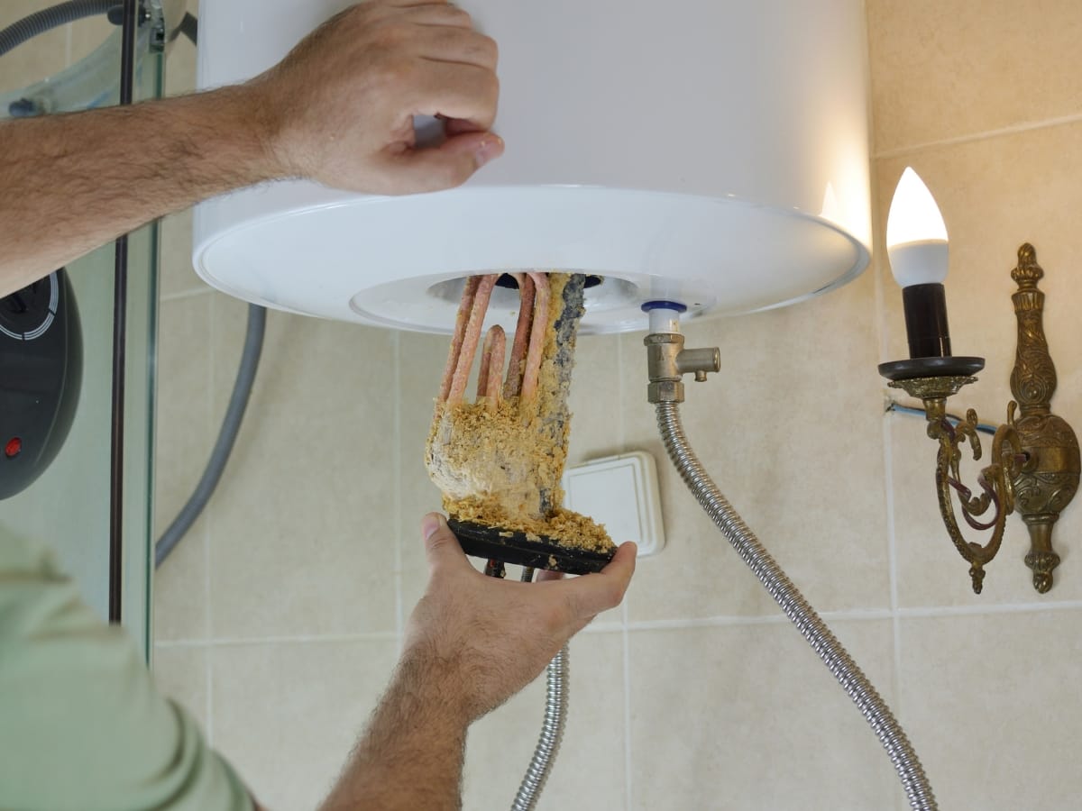 A person is cleaning a water heater element covered in mineral scale inside a bathroom with tiled walls, highlighting the importance of maintenance similar to an annual furnace inspection. A wall-mounted light fixture is nearby.